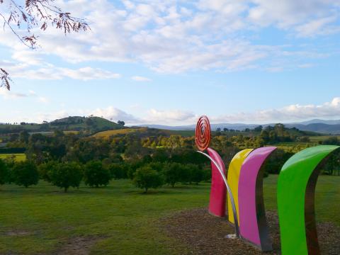 Rolling green hills studded with trees, near sunset. An abstract sculpture in bright glossy colours in the foreground.